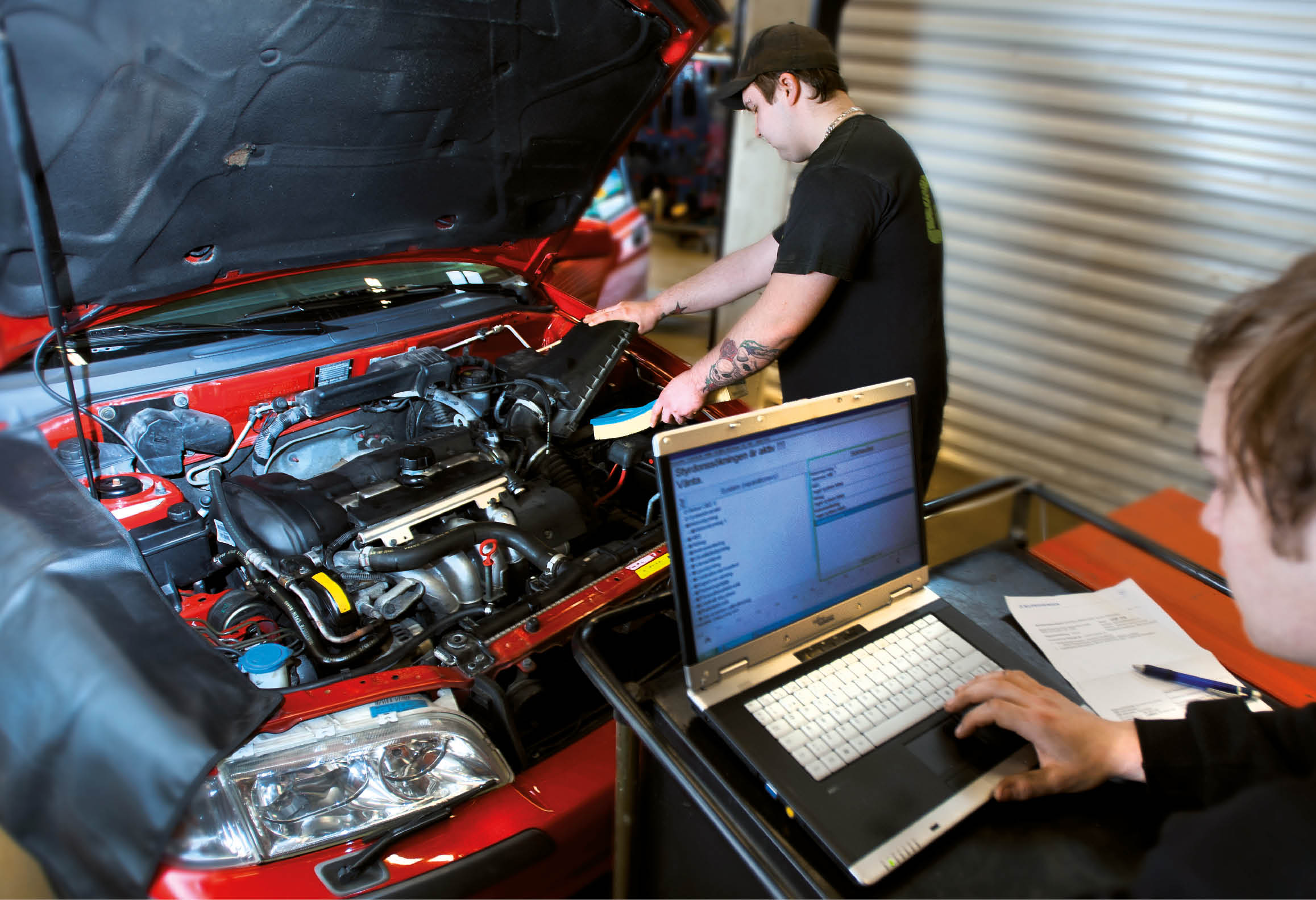 En elev arbetar med bilmotor och annan elev använder laptop 